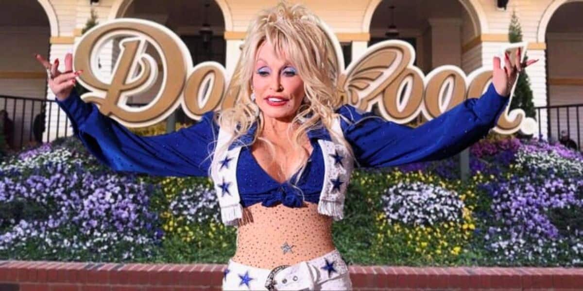 A person with long blonde hair and a sparkly blue and white outfit standing in front of the Dollywood sign, channeling their inner Dolly Parton. They are smiling with their arms raised. The background includes colorful flowers and part of a building.