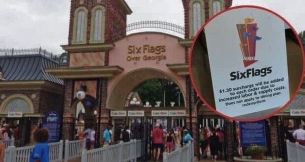 Entrance of Six Flags Over Georgia theme park with a sign in the foreground noting a $1.30 surcharge for orders due to increased labor and supply costs. People are visible near the gates.