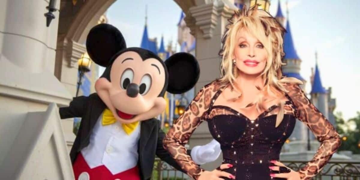 In front of a castle backdrop, a person dressed as Mickey Mouse stands next to a woman with blonde hair wearing a black dress with leopard print sleeves. Both are smiling and posing for the camera.