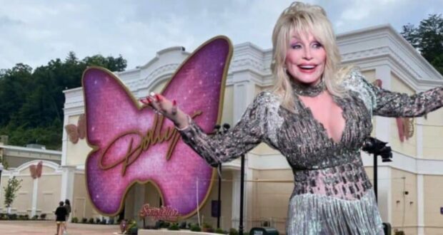 A woman with blonde hair and wearing a glittery silver dress poses with arms outstretched in front of a white building. The building has a large pink butterfly sign with the text "Dollywood" on it. Trees and hills are visible in the background.