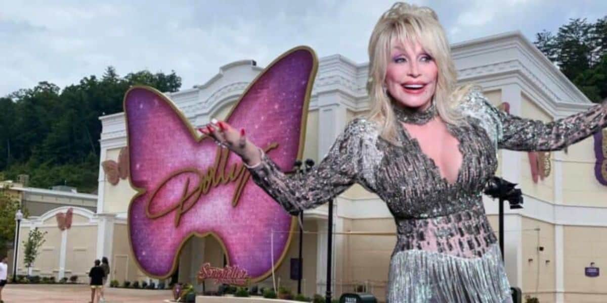 A woman with blonde hair and wearing a glittery silver dress poses with arms outstretched in front of a white building. The building has a large pink butterfly sign with the text "Dollywood" on it. Trees and hills are visible in the background.