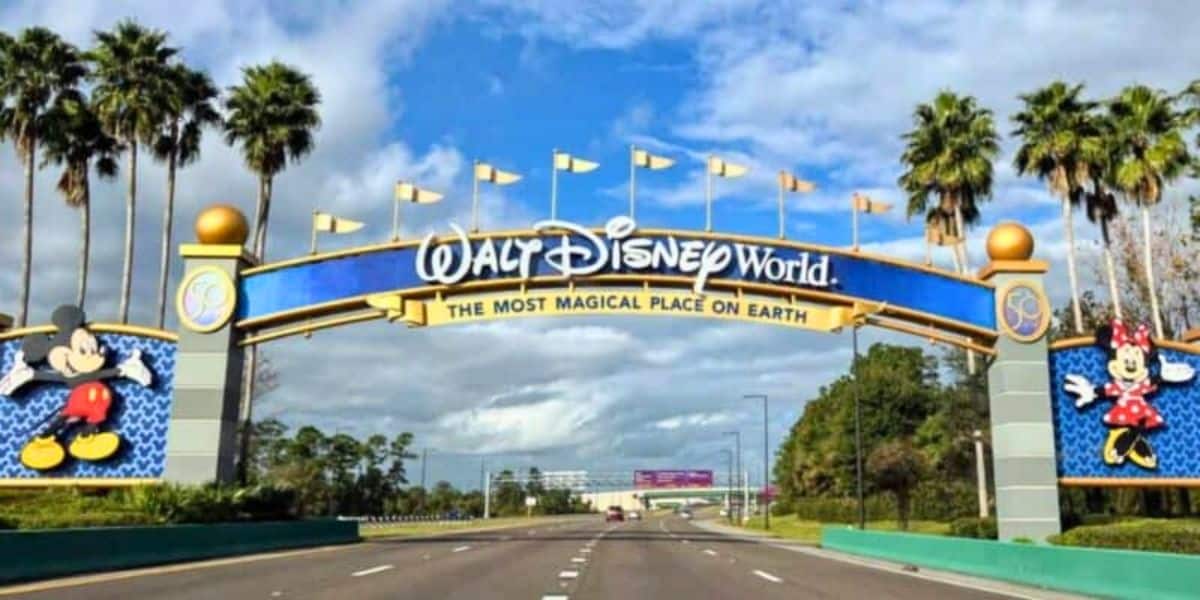 Entrance sign of Walt Disney World, featuring large bold letters and images of Mickey and Minnie Mouse, under a sunny sky with fluffy clouds, viewed from a road lined with palm trees near Typhoon