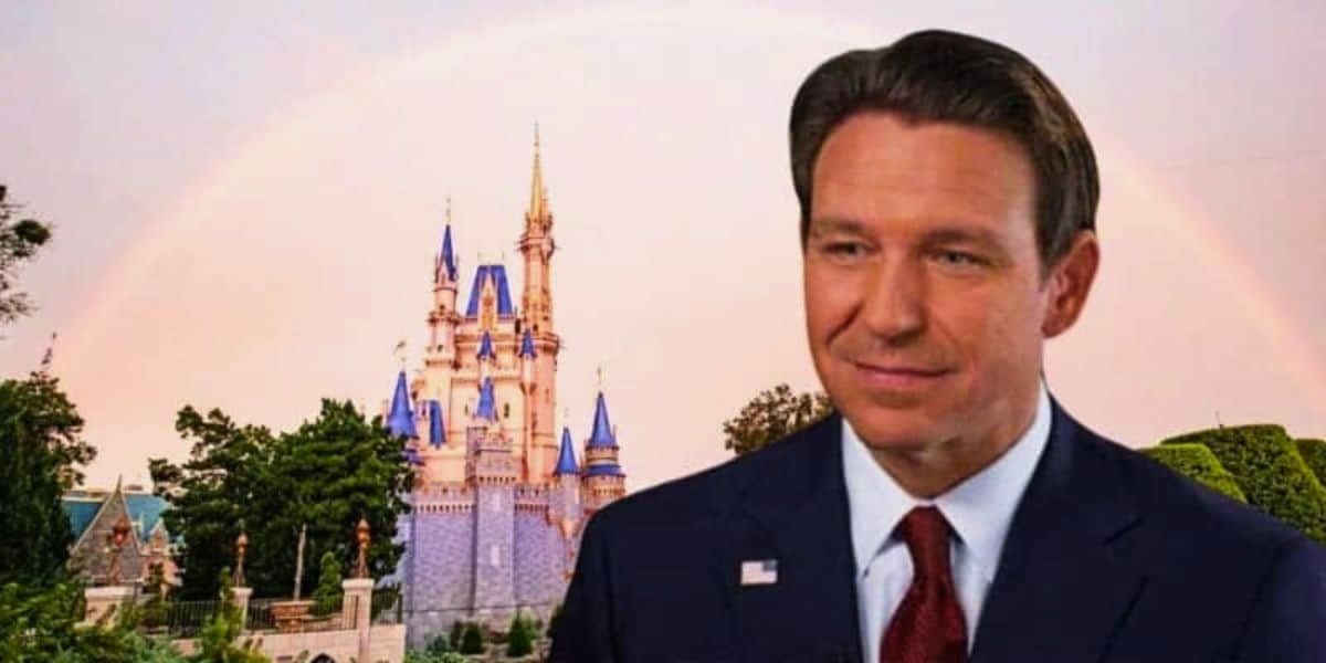 A man in a blue suit with a red tie stands superimposed on an image of disneyland featuring sleeping beauty's castle under a bright rainbow.