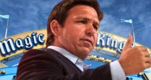 Ron DeSantis giving a thumbs up in front of the Magic Kingdom entrance sign under a blue sky.
