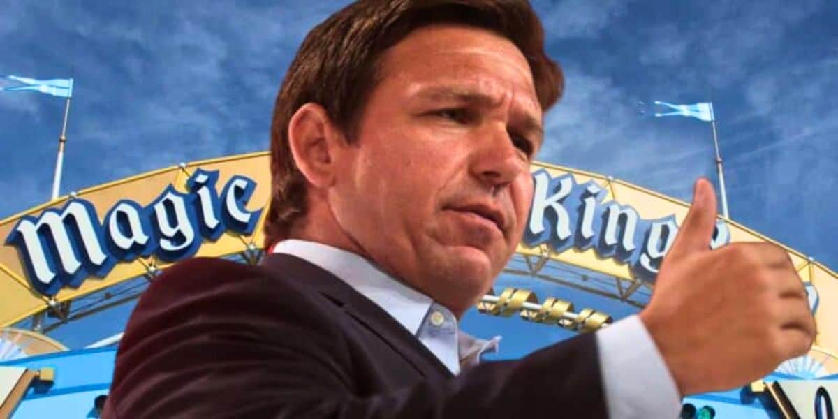 Ron DeSantis giving a thumbs up in front of the Magic Kingdom entrance sign under a blue sky.
