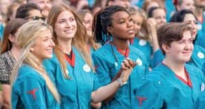 A diverse group of people, mostly young adults, are standing closely together, smiling, and looking ahead. They are all wearing teal uniforms with badges. Some are holding hands, creating a sense of unity and celebration. The background shows more people in similar attire.