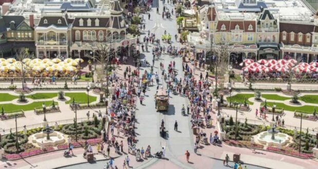 A busy, bird's-eye view of a vibrant theme park with numerous visitors walking around landscaped gardens, patterned walkways, and colorful umbrellas dotting the area.