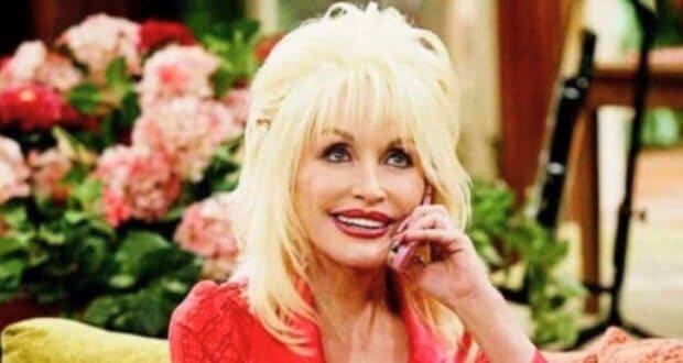 Dolly Parton with voluminous hair, wearing a bright red top, sits on a couch smiling and holding a cellphone to her ear. Behind her, there are pink flowers and lush greenery, creating a warm and inviting atmosphere.