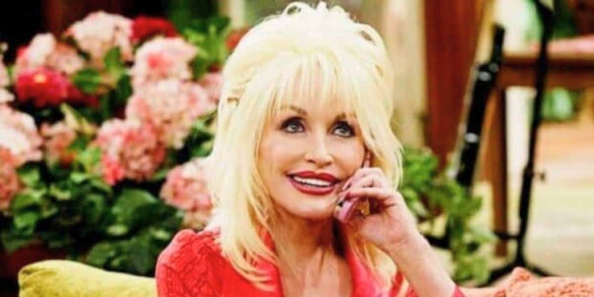 Dolly Parton with voluminous hair, wearing a bright red top, sits on a couch smiling and holding a cellphone to her ear. Behind her, there are pink flowers and lush greenery, creating a warm and inviting atmosphere.
