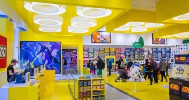 A vibrant LEGO store with bright yellow walls and ceiling invites enthusiasts to explore. Shoppers admire displays featuring various sets, including a standout R2-D2 model in the Star Wars section. Fans gather eagerly around diverse product areas, sharing their passion for the LEGO universe.