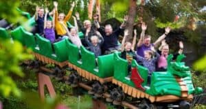 A group of people are enjoying a ride on a green dragon-themed roller coaster at the theme park. They have their hands up and are smiling and laughing. The coaster is surrounded by lush green trees and foliage, suggesting a lively and fun atmosphere at the amusement park.