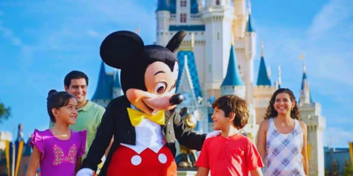 A family of four beams with joy standing next to a cheerful Mickey Mouse at a magical theme park. Behind them, a majestic castle with blue spires rises against the clear sky, capturing their excitement and happiness.