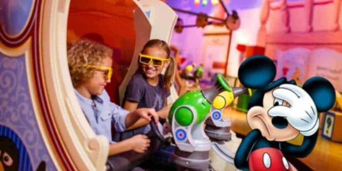 Two children seated in a Disneyland ride vehicle with 3D glasses, smiling and holding blasters. A whimsical backdrop and colorful lighting enhance the amusement park setting. Meanwhile, a life-sized Mickey Mouse figure stands nearby, playfully covering his face with one hand, perhaps bashfully avoiding the viral hack that's been criticized for destroying Disney property.