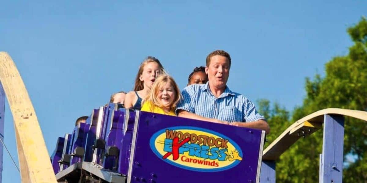A group of people, including a man in a blue plaid shirt and a young girl in a yellow shirt, enjoy a thrilling ride on the Woodstock Express roller coaster at Carowinds Theme Park. They look excited and joyful as the coaster descends a hill under a clear blue sky.
