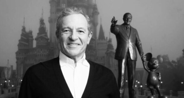A man smiles broadly while standing in front of a castle and a statue of a man holding hands with a mouse character, iconic symbols of the Disney Company. The image is in black and white, and the atmosphere appears slightly foggy.
