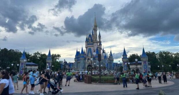 A bustling crowd gathers in front of a large, ornate castle with blue spires and golden accents under a partly cloudy sky at an amusement park. Paths are lined with lampposts and foliage, with visitors walking, pushing strollers, and taking photos.
