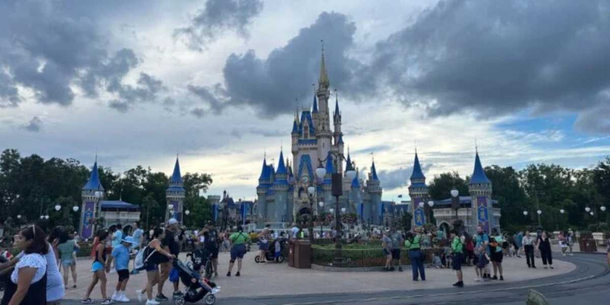 A bustling crowd gathers in front of a large, ornate castle with blue spires and golden accents under a partly cloudy sky at an amusement park. Paths are lined with lampposts and foliage, with visitors walking, pushing strollers, and taking photos.