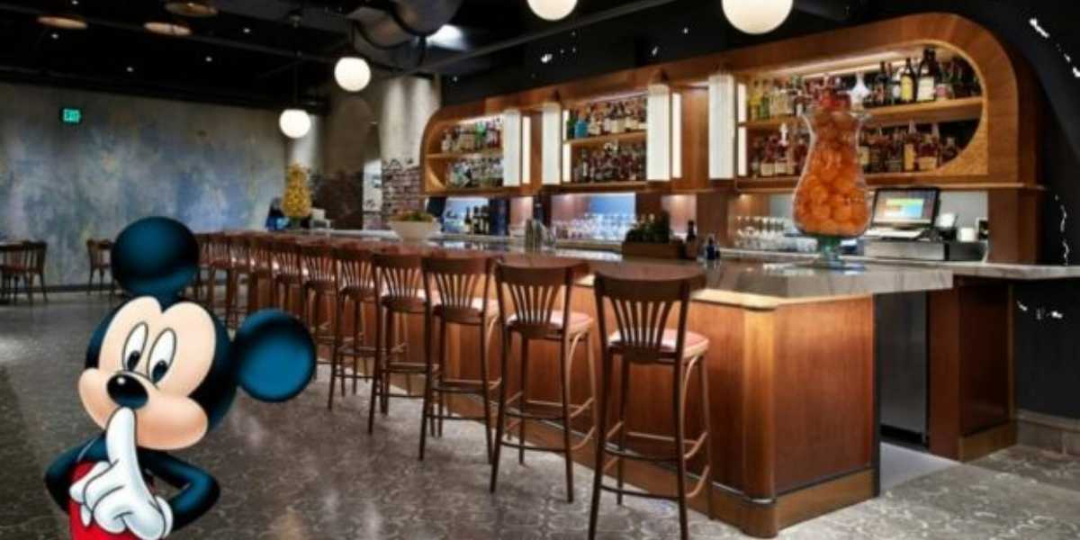 A modern bar with wooden chairs and a marble countertop is shown. Various bottles and glasses are displayed on shelves behind the bar. A figure of Mickey Mouse is in the foreground, holding a finger to his lips as if signaling to be quiet, capturing the secretive vibe of Enzo's Hideaway.