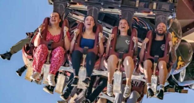 Four people are seated on an amusement park ride, suspended upside-down. They appear to be experiencing a range of emotions from excitement to fear. The bright, clear blue sky serves as the background.