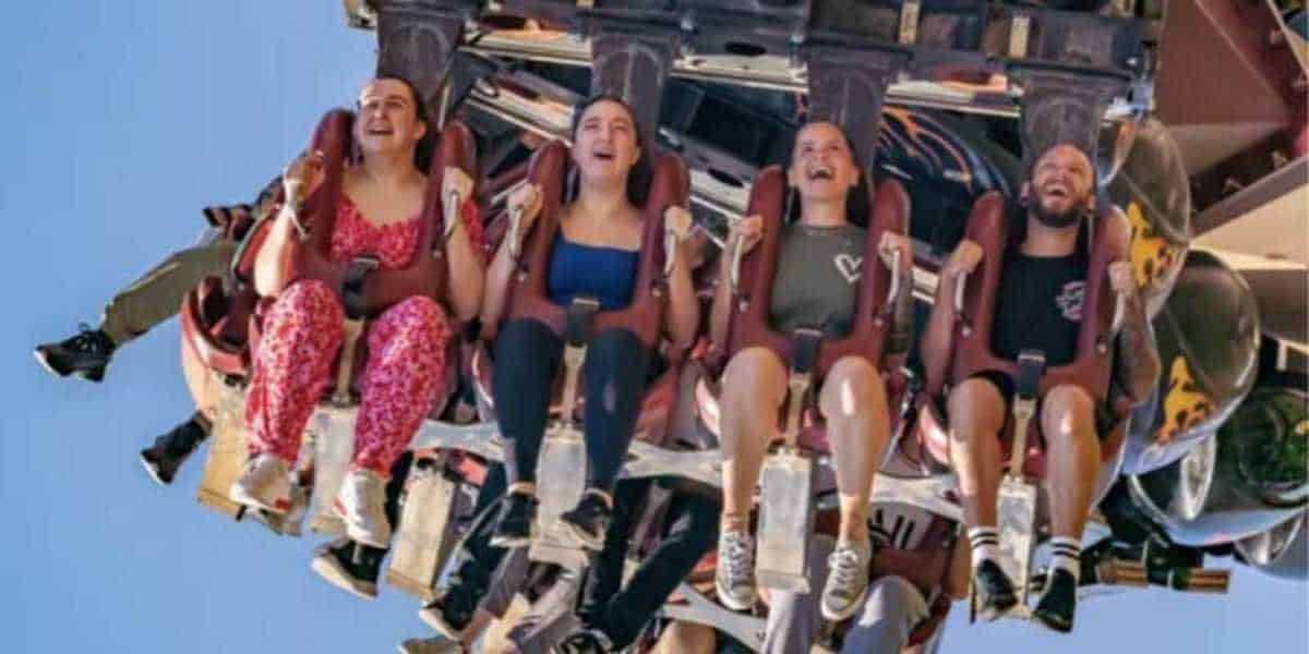 Four people are seated on an amusement park ride, suspended upside-down. They appear to be experiencing a range of emotions from excitement to fear. The bright, clear blue sky serves as the background.