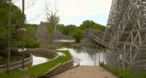 An abandoned amusement park, the remnants of a theme park shut down, featuring a wooden roller coaster partially submerged in floodwater. Overgrown vegetation surrounds the area. The sky is overcast, and a fenced-off path leads toward the flooded section. A red hose is laid on the path.