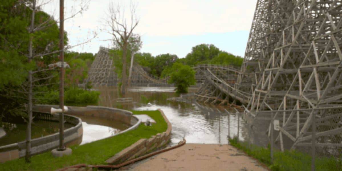 An abandoned amusement park, the remnants of a theme park shut down, featuring a wooden roller coaster partially submerged in floodwater. Overgrown vegetation surrounds the area. The sky is overcast, and a fenced-off path leads toward the flooded section. A red hose is laid on the path.