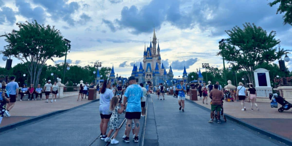 Visitors walk down a broad pathway towards a grand, iconic castle with blue and gold accents. The sky is partly cloudy, and trees line the pathway. People are taking photos, chatting, and enjoying the theme park atmosphere.