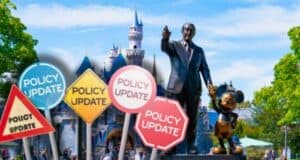 A statue of Walt Disney holding hands with a Mickey Mouse stands in front of a castle at a Disneyland Park in California. In the foreground, various road signs with "Policy Update" written on them are visible. The sky is clear and trees frame the image on the sides.