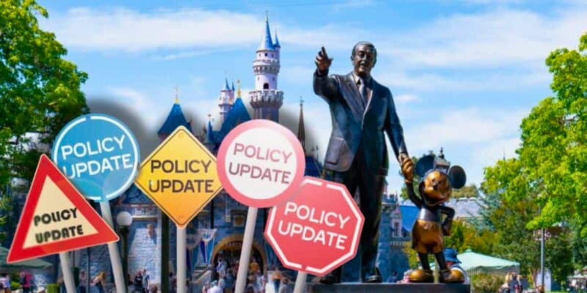 A statue of Walt Disney holding hands with a Mickey Mouse stands in front of a castle at a Disneyland Park in California. In the foreground, various road signs with "Policy Update" written on them are visible. The sky is clear and trees frame the image on the sides.