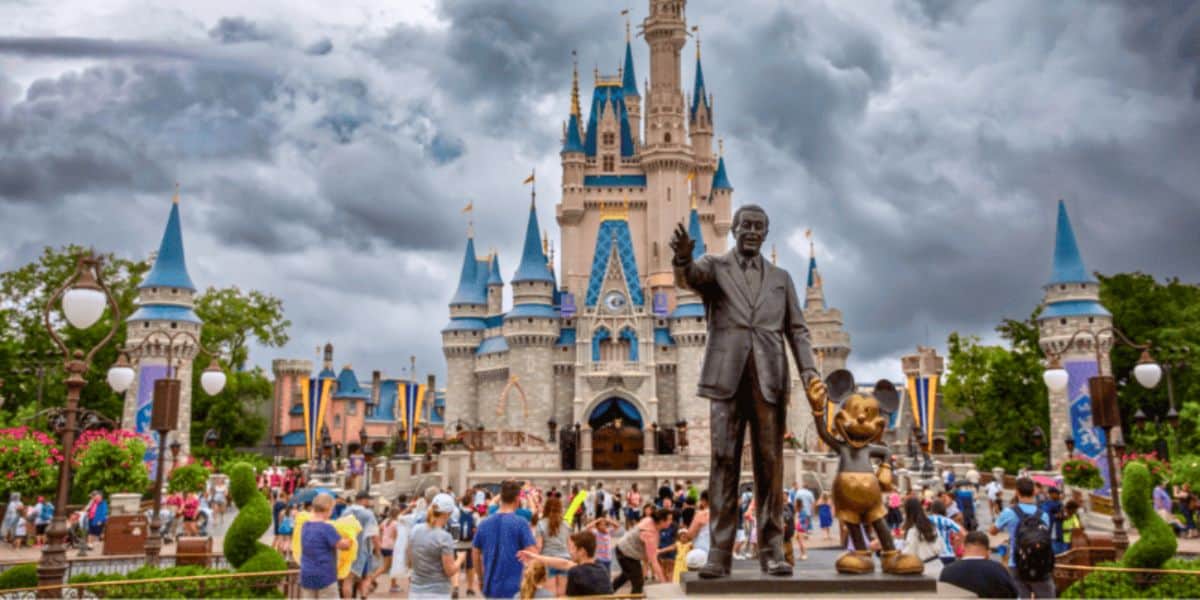 A vibrant scene at a theme park featuring a large castle with blue turrets under a cloudy sky. In the foreground, a statue of a person holding hands with a cartoon mouse overlooks a bustling crowd of visitors. Lush greenery and flags decorate the surroundings.