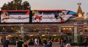 A monorail adorned with Disney characters, including Goofy and Mickey Mouse, moves along an elevated track. People walk below the track, and the entrance to Disneyland with its recognizable sign and architecture is visible in the background, illustrating a magical world despite transportation cuts.