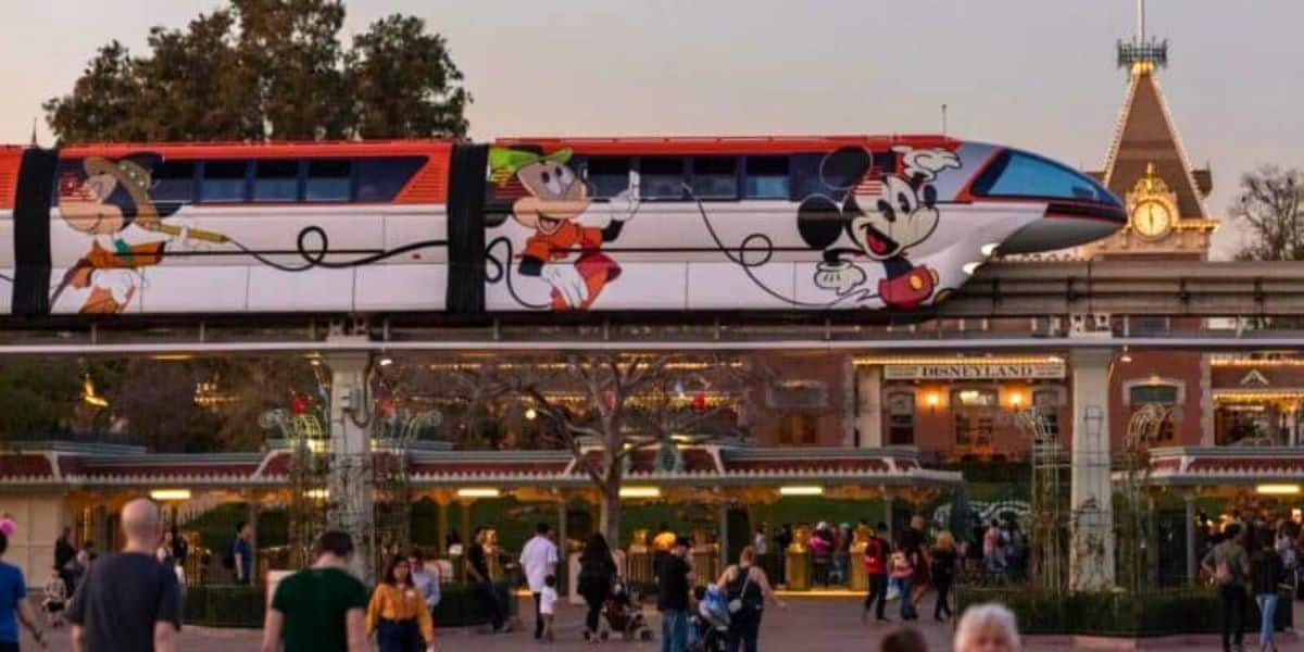 A monorail adorned with Disney characters, including Goofy and Mickey Mouse, moves along an elevated track. People walk below the track, and the entrance to Disneyland with its recognizable sign and architecture is visible in the background, illustrating a magical world despite transportation cuts.