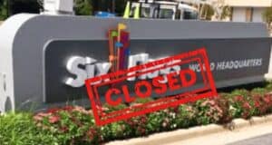 A sign indicating the corporate headquarters of Six Flags, with the words "WORLD HEADQUARTERS" visible. A bold red "CLOSED" stamp is overlaid on the image. The sign is surrounded by plants and shrubs, giving a shuttered look to the once-busy location.