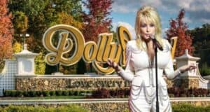 A performer in a white, embellished outfit stands in front of a sign that reads "Dollywood" in an amusement park setting, surrounded by autumn foliage. They hold a microphone and gesture with one hand. The backdrop includes fall-colored trees and a sunny sky, echoing the charm of Dolly Parton's vibrant vision.