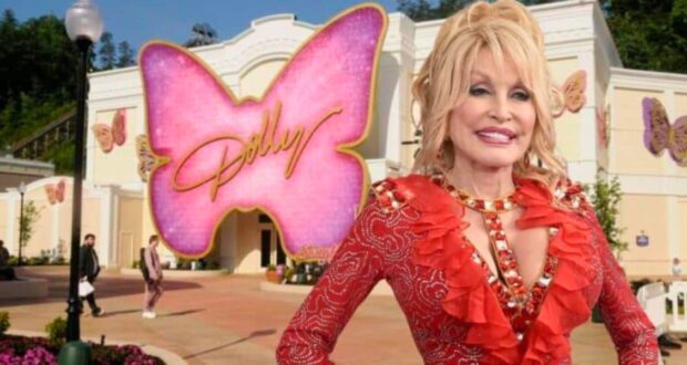 A cheerful woman with blonde hair wearing a red, jeweled outfit stands in front of a building decorated with large pink and beige butterfly designs. The largest pink butterfly in the background has the word “Dolly” written on it. People are walking around the area.