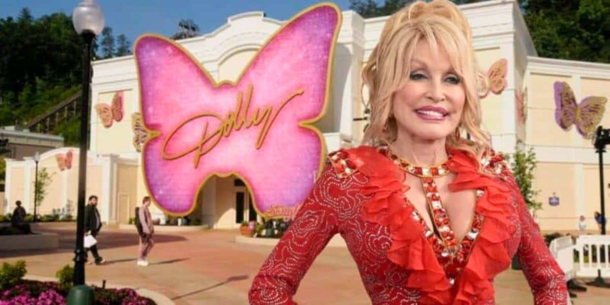 A cheerful woman with blonde hair wearing a red, jeweled outfit stands in front of a building decorated with large pink and beige butterfly designs. The largest pink butterfly in the background has the word “Dolly” written on it. People are walking around the area.