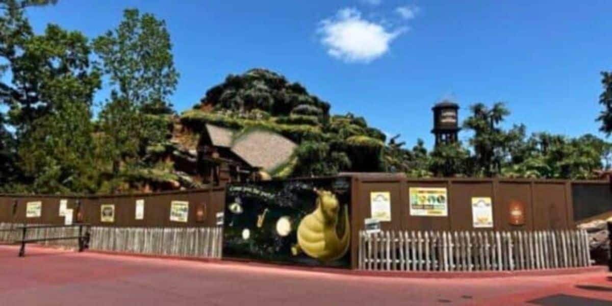 A large construction wall surrounds Tiana's Bayou Adventure, a mountain with greenery and a rocky hill reminiscent of Splash Mountain visible in the background.