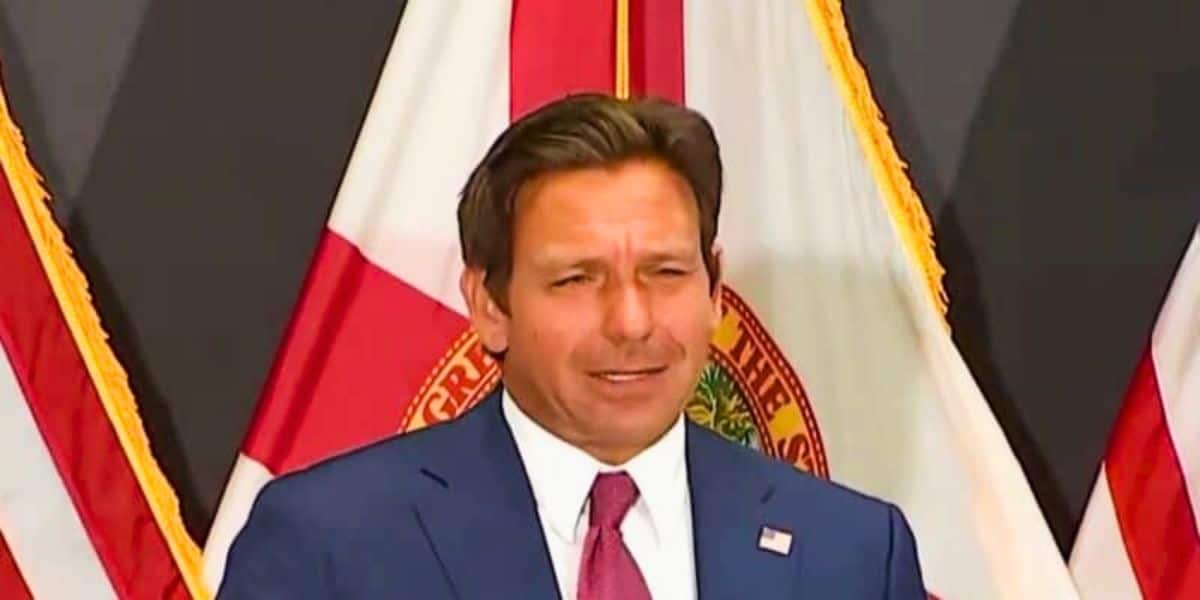 Ron DeSantis is standing in front of a backdrop featuring red and white flags. He has short brown hair and is speaking or making a gesture with his right hand.
