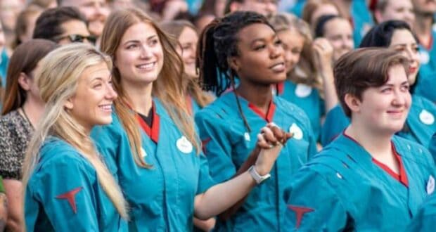 A diverse group of Disney cast members, mostly young adults, are standing closely together, smiling, and looking ahead. They are all wearing teal uniforms with badges. Some are holding hands, creating a sense of unity and celebration. The background shows more people in similar attire.