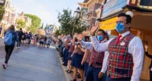 A group of Disneyland cast members, dressed in red and blue plaid vests and blue face masks, line both sides of a street, waving to guests who are walking towards the camera.