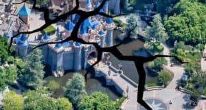 An aerial view of a theme park castle with blue rooftops and surrounding gardens, featuring large, dark cracks superimposed over the image, giving the appearance of a shattered or broken visual effect. The scene includes lush greenery and pathways.
