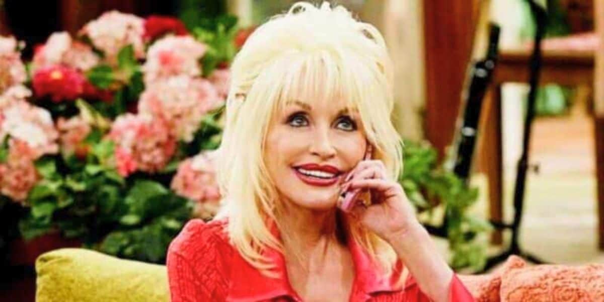 Dolly Parton with blond hair and a bright smile is sitting on a couch, holding a pink phone to her ear. Behind her, there are colorful flowers and green plants in a cozy indoor setting.