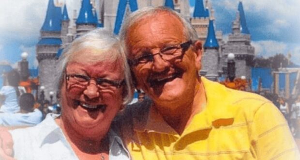 An elderly couple, smiling warmly, pose in front of a castle with blue turrets, likely at an amusement park. The woman has short white hair and wears glasses and a white shirt, while the man, also with glasses, wears a yellow polo shirt.