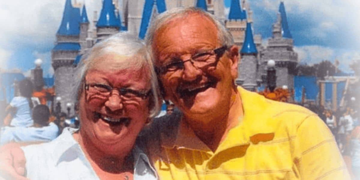 An elderly couple, smiling warmly, pose in front of a castle with blue turrets, likely at an amusement park. The woman has short white hair and wears glasses and a white shirt, while the man, also with glasses, wears a yellow polo shirt.