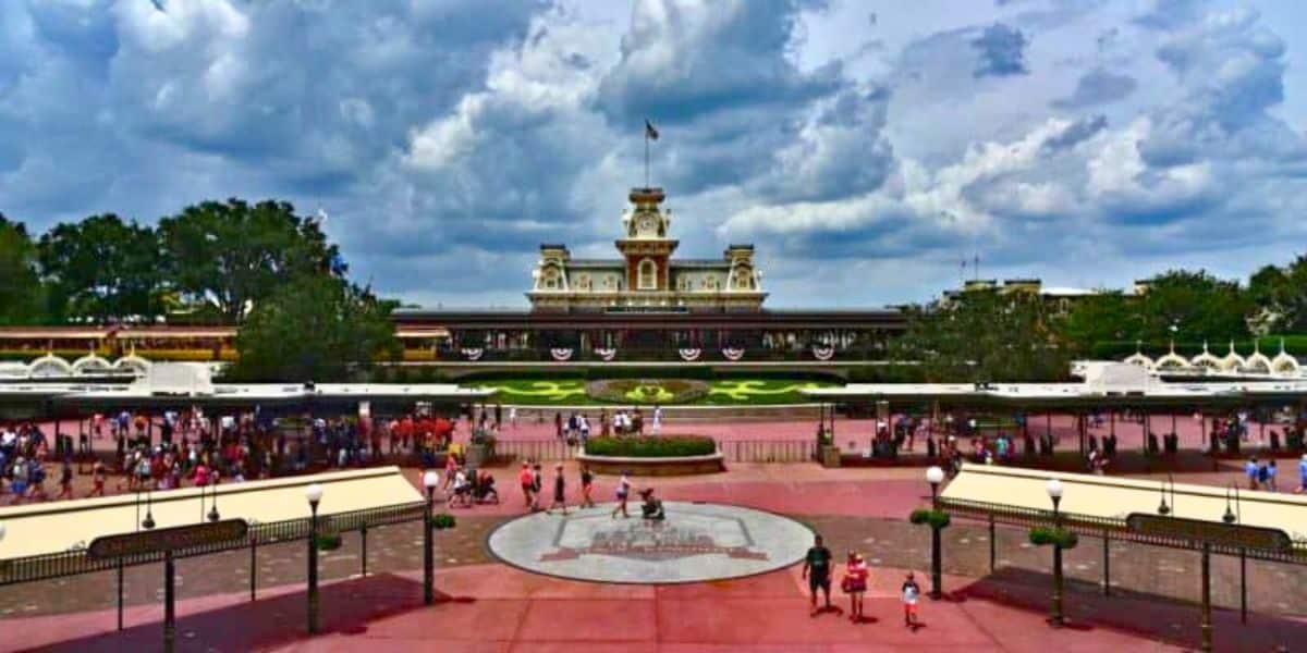 View of the entrance to Disney World, featuring a railway station in the background, bustling with visitors under a cloudy sky.