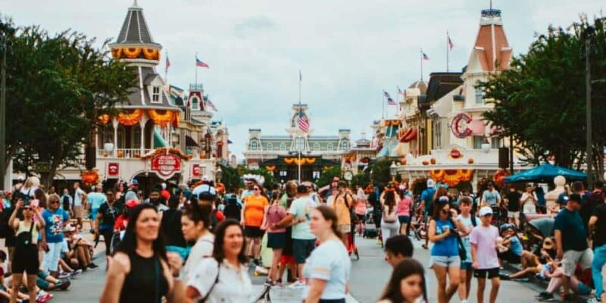A bustling theme park scene unfolds along Main Street America, with a crowd of people walking past buildings adorned with festive bunting and American flags. Some visitors rest on the ground, while a cloudy sky looms overhead.