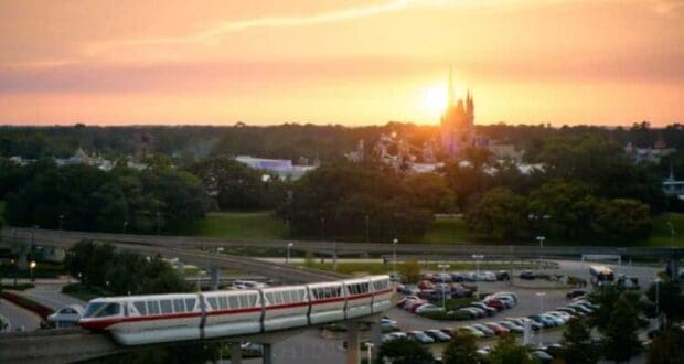 A monorail travels along elevated tracks in the foreground, with a vast parking lot below. In the background, trees surround a castle-like structure, and the sky is illuminated by a vibrant sunset.