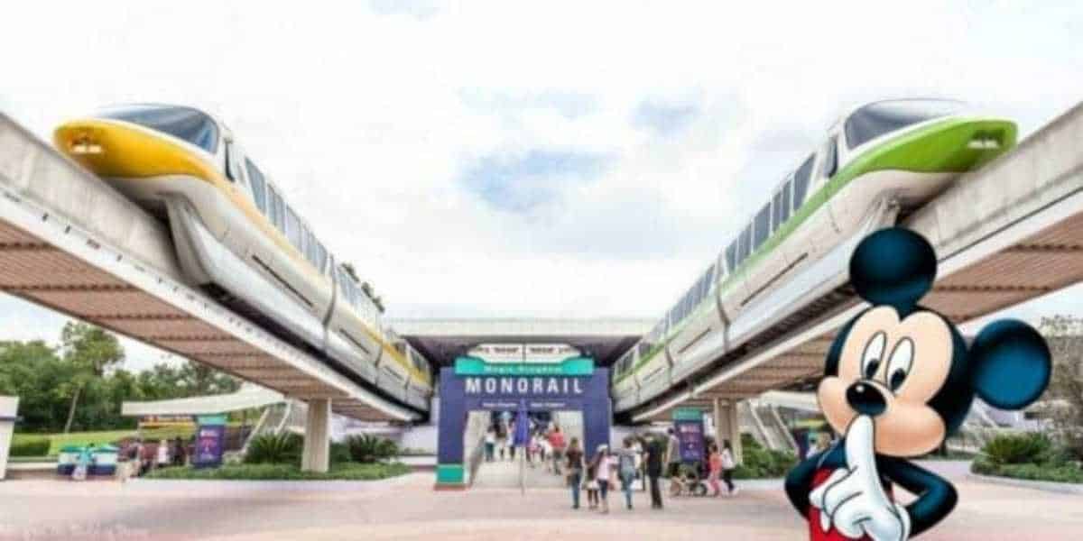 Two monorail trains, one yellow and one green, are positioned on parallel elevated tracks. A crowd stands below near a sign that reads "Monorail." In the foreground, Mickey Mouse is shown in a "shh" gesture, perhaps hinting at a Disney Transportation and Ticket Center trick.