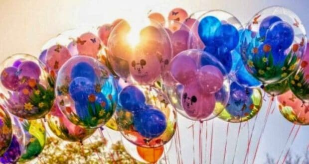 A cluster of colorful balloons floats against a bright, sunny sky. The balloons are inside clear outer balloons with vibrant designs, including flowers, butterflies, and cartoon faces, creating a whimsical and joyful scene. In the background, you can slightly see trees as a dad refuses to take his daughter to Disney.