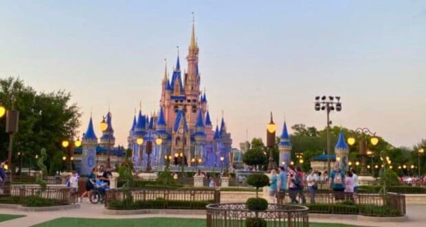 A view of Cinderella Castle at dusk in Walt Disney World Free. The castle is illuminated with blue and gold lights, surrounded by people. Trees and lampposts are visible, with the sky gradually transitioning to night. The scene captures a moment of magic and wonder.